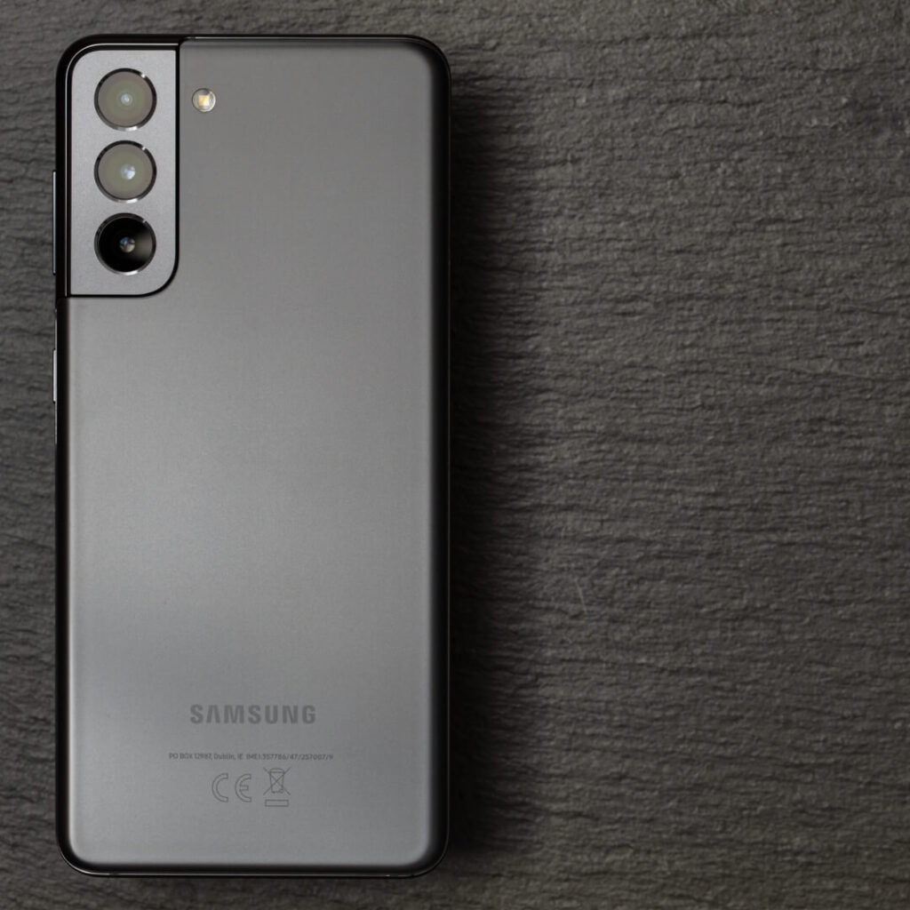 A Samsung smartphone with a triple rear camera setup is placed on a textured dark surface, shown from the back.