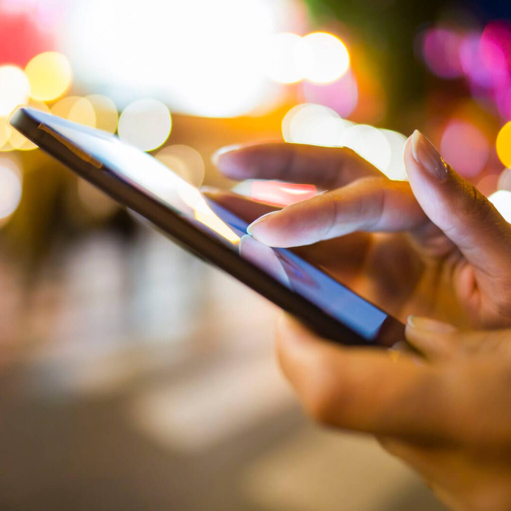 Close-up of a person’s hand using a smartphone touchscreen outdoors at night, with colourful blurred city lights in the background.
