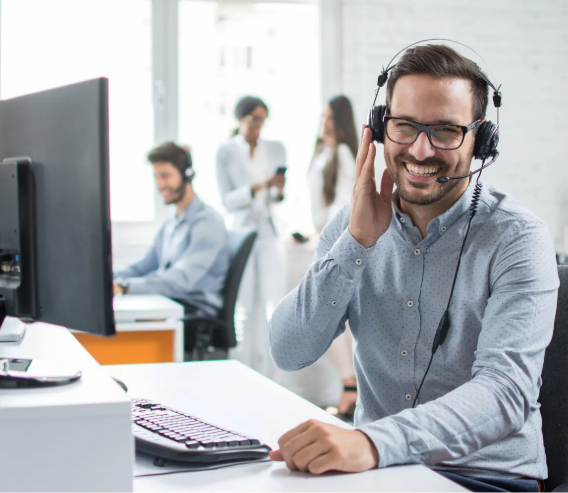 Man with headset smiling at desk, colleagues talking in background, office setting with computers.