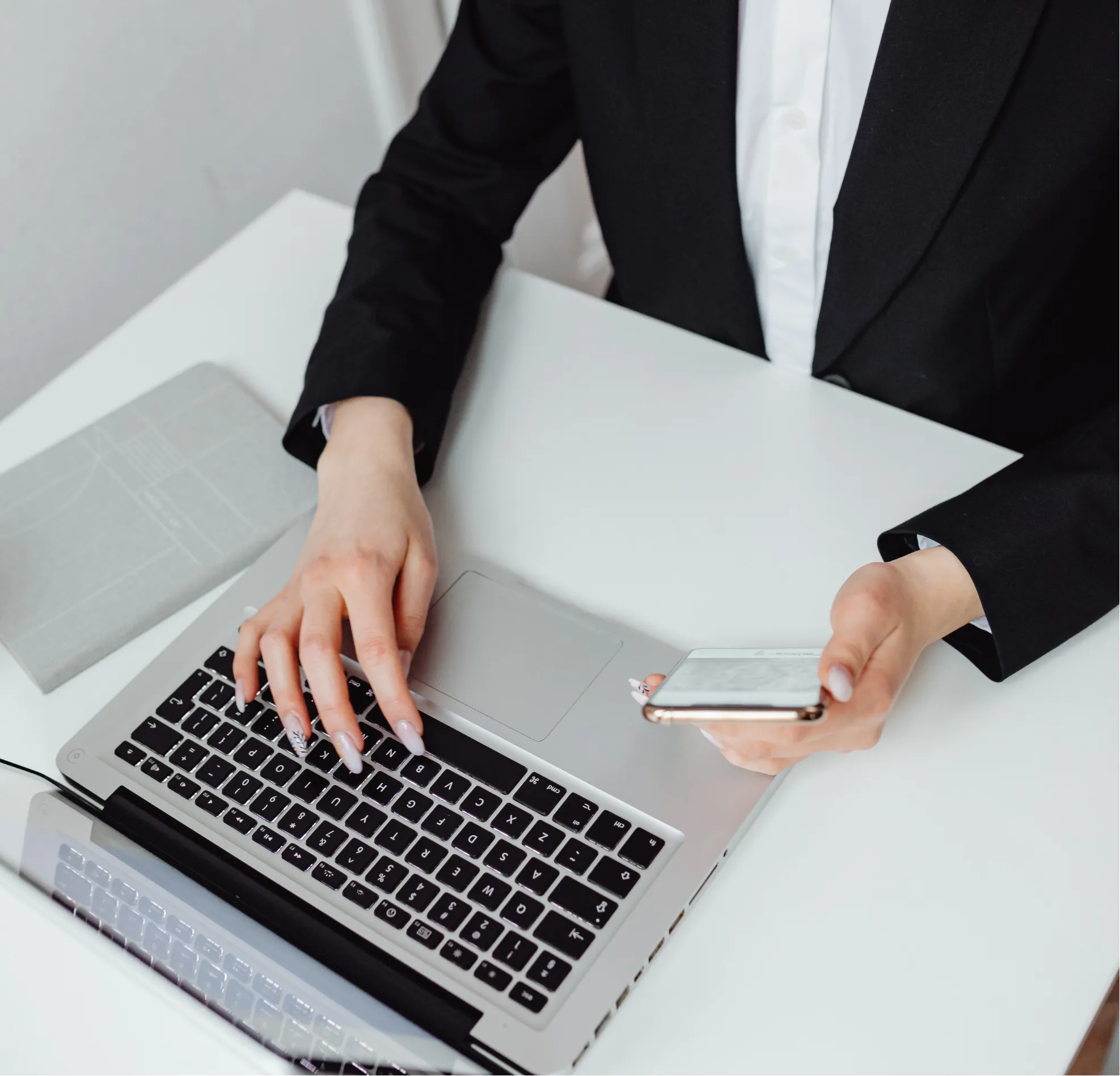 Person in a suit using a laptop and holding a mobile phone at a white desk.