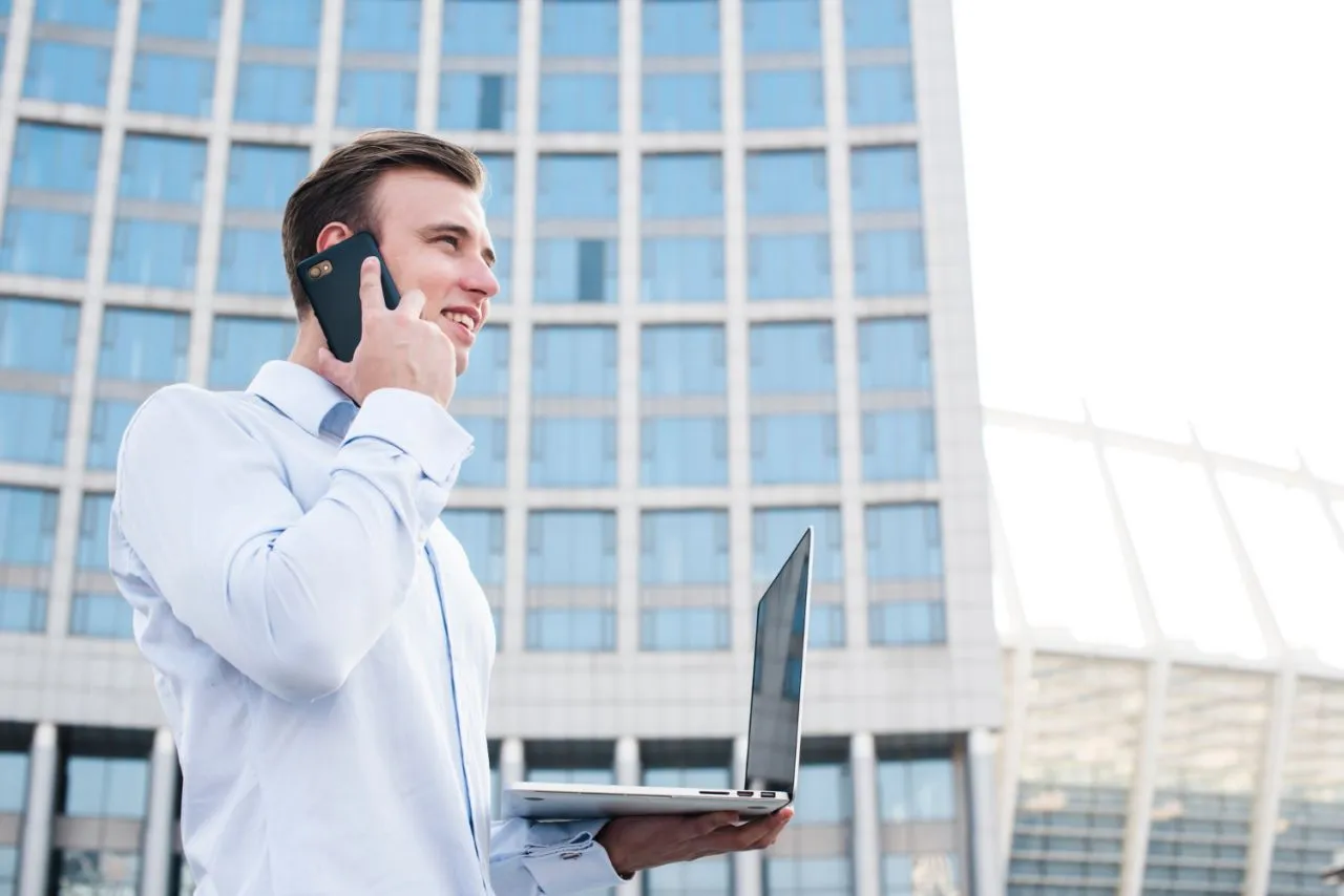 A smiling man in a white shirt holds a laptop and talks on the phone outside a glass office block.